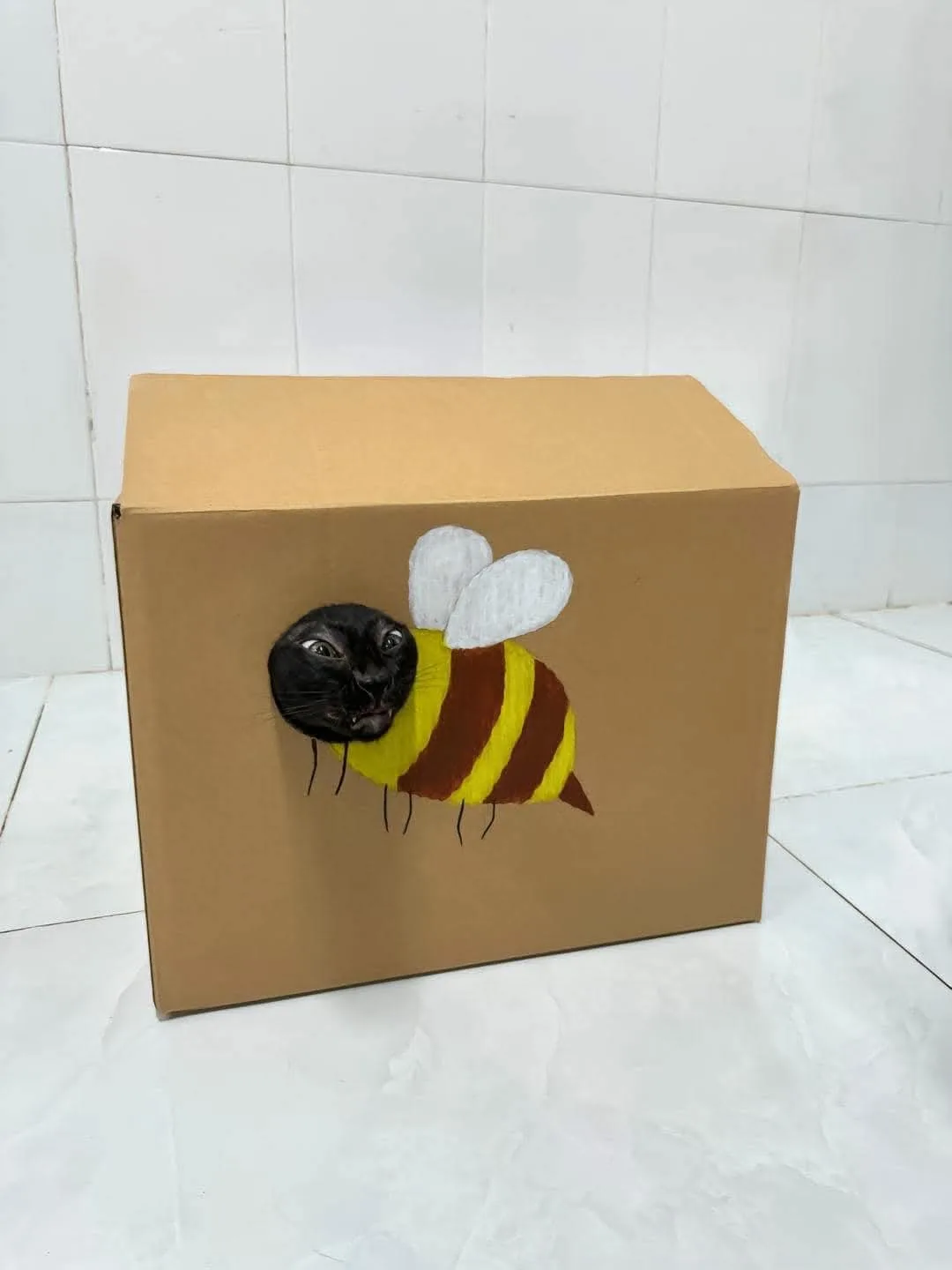 A black cat peeks out from a cardboard box that has been painted to look like a bee, complete with yellow and brown stripes…