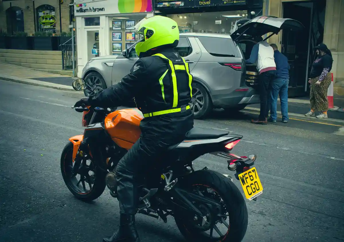A motorcyclist in a bright yellow helmet and black gear with neon yellow reflective stripes sits on an orange Honda…