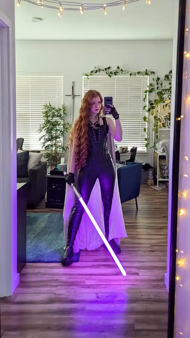 A person with long red hair takes a mirror selfie in a living room, dressed in a black leather outfit with a white cape and…