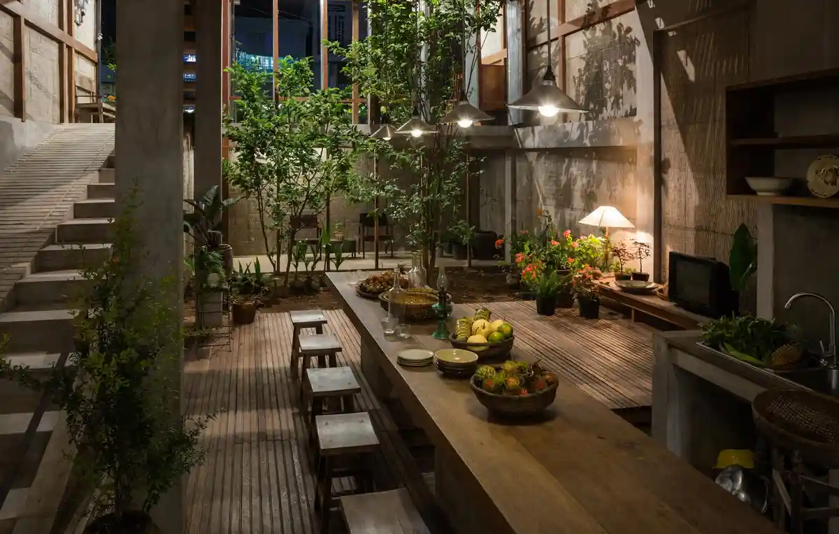 A warmly lit, rustic indoor space features a long wooden counter with stools, bowls of tropical fruit, potted plants…