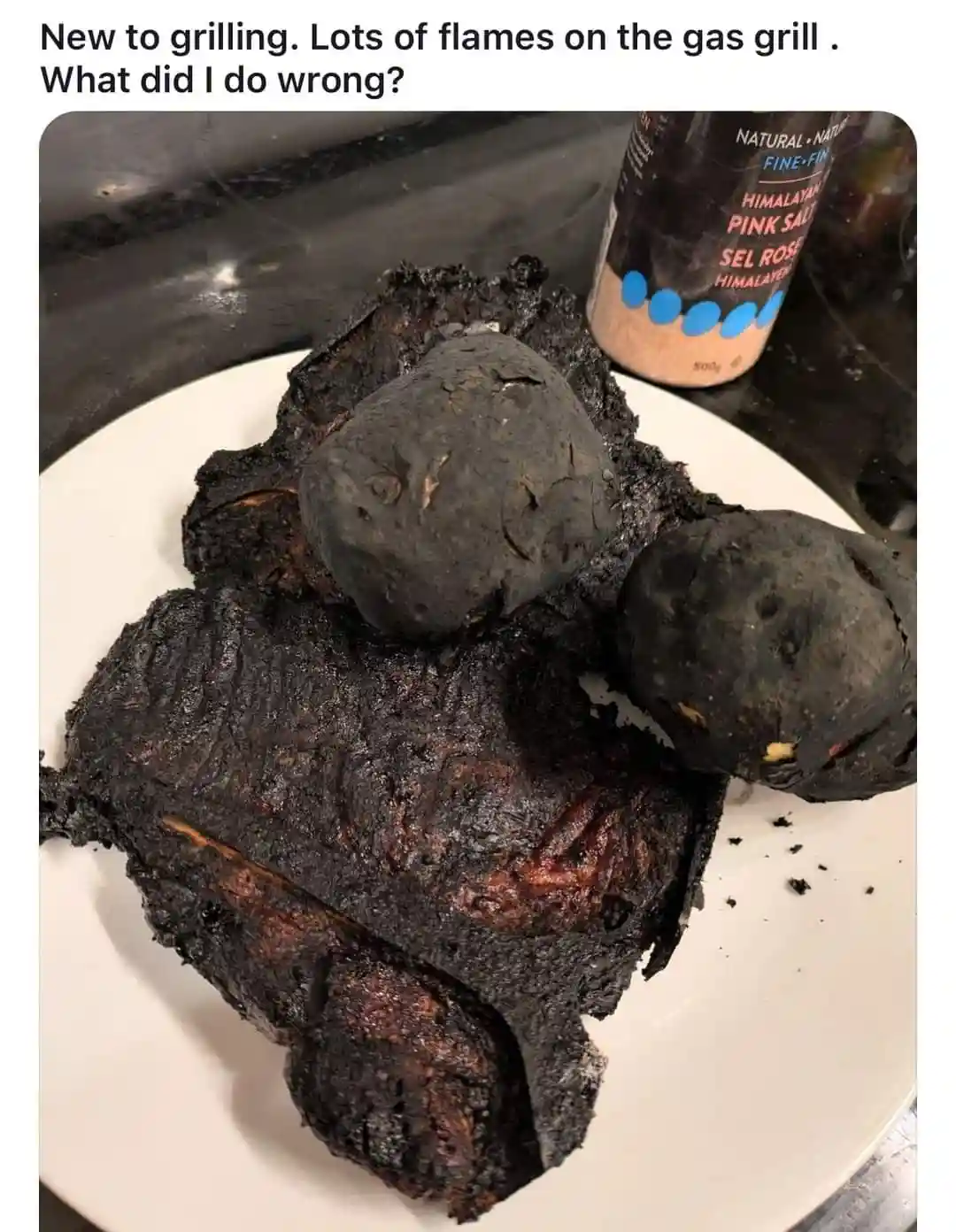 A white plate holds severely charred, blackened food items that look like burnt meat and potatoes, with a bottle of…