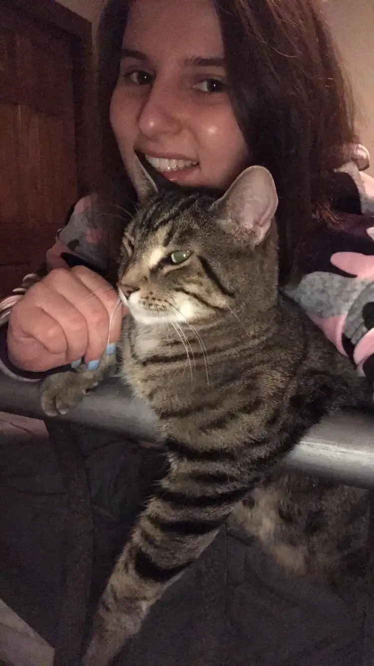 A young woman with dark hair smiles broadly while holding a tabby cat that looks unimpressed and is staring off to the side…