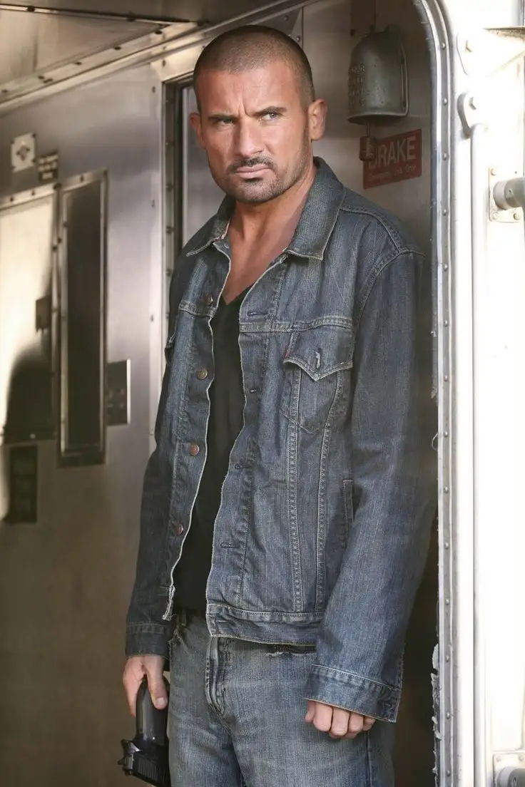 A man with a shaved head and goatee, wearing a denim jacket and jeans, stands in the doorway of a train car holding a…