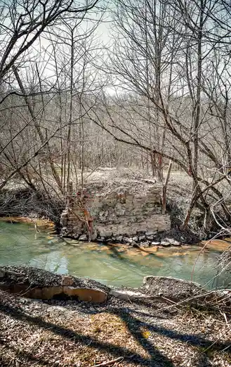 A photograph of a dilapidated stone structure partially submerged in a shallow, murky river, surrounded by bare winter…