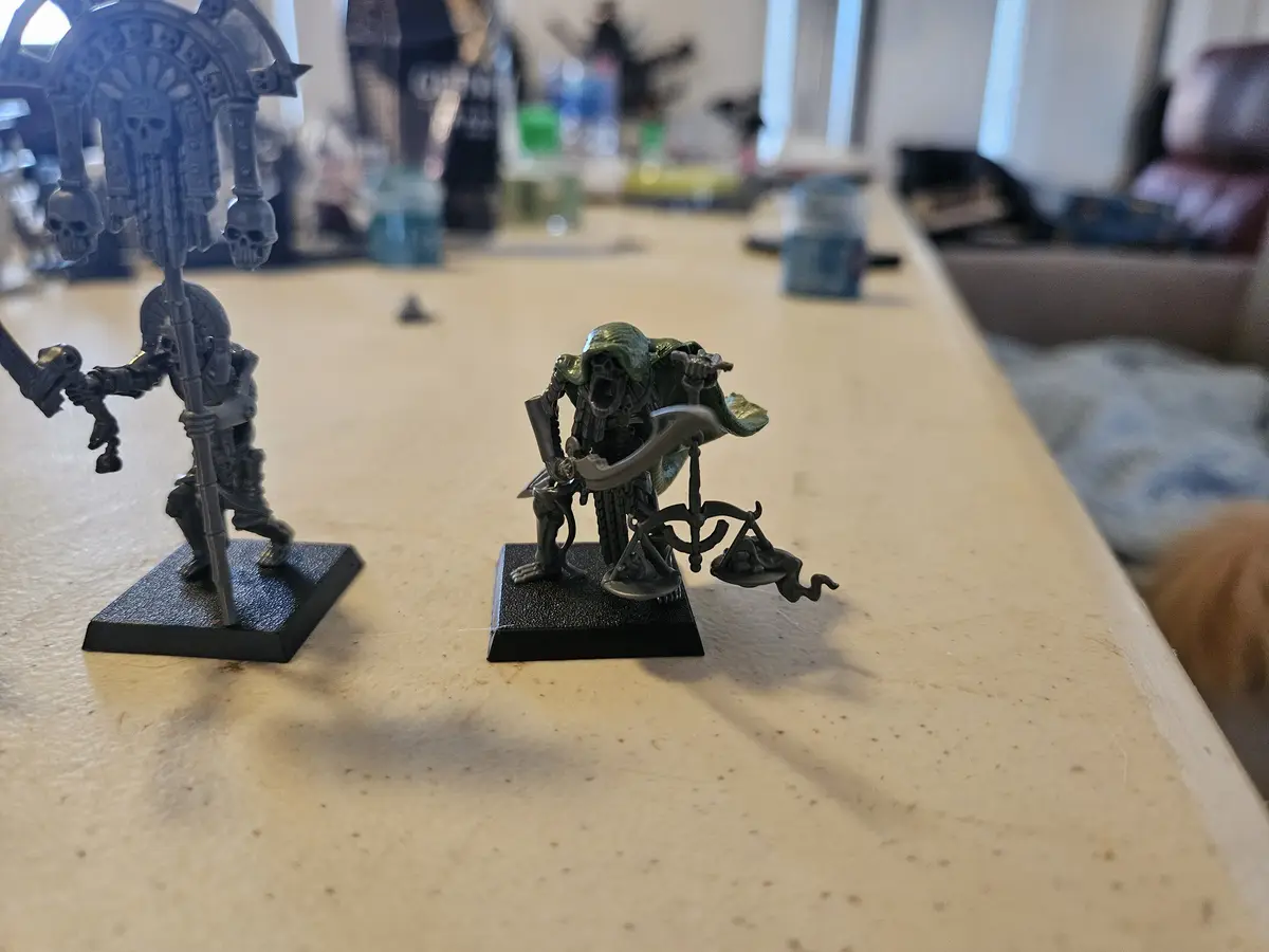 Two painted miniature figurines sit on a table; one is a grey, armored warrior holding a staff with skulls, the other is a…