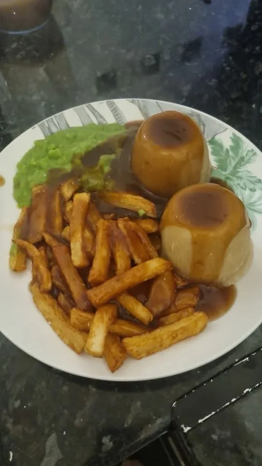 a plate of food featuring two round caramelized puddings, french fries, and green mushy peas, all covered in gravy. The text…