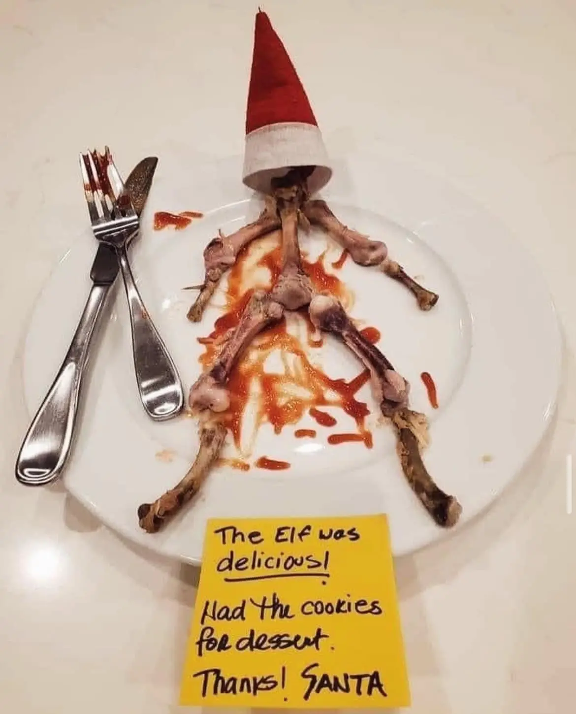 A white plate holds leftover chicken bones arranged to resemble a human figure wearing a red and white Santa hat, with sauce…