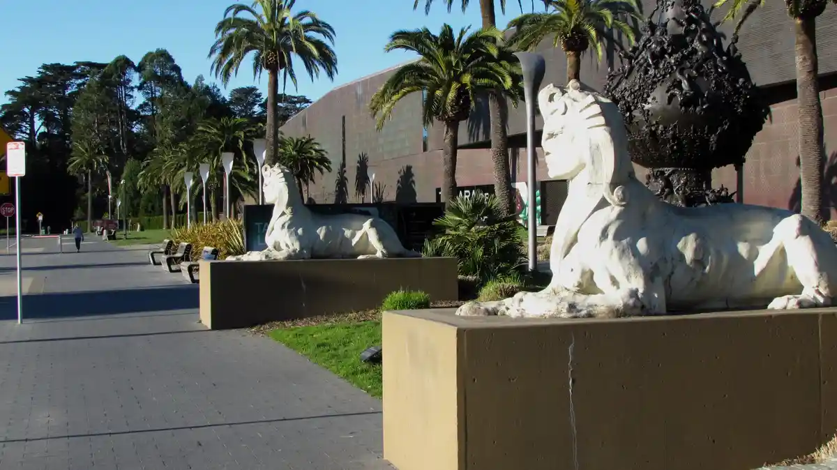 Two large, white, stone sphinx statues flank a paved walkway outside a modern building with palm trees. The sphinx on the…