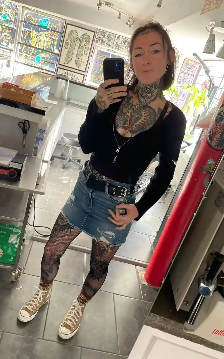 A woman with extensive tattoos covering her neck, chest, arms, and legs stands in a tattoo parlor, taking a mirror selfie…