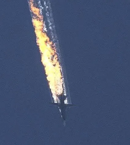 A dark-colored jet aircraft is seen flying against a clear blue sky, trailing a thick, bright orange and yellow flame and…