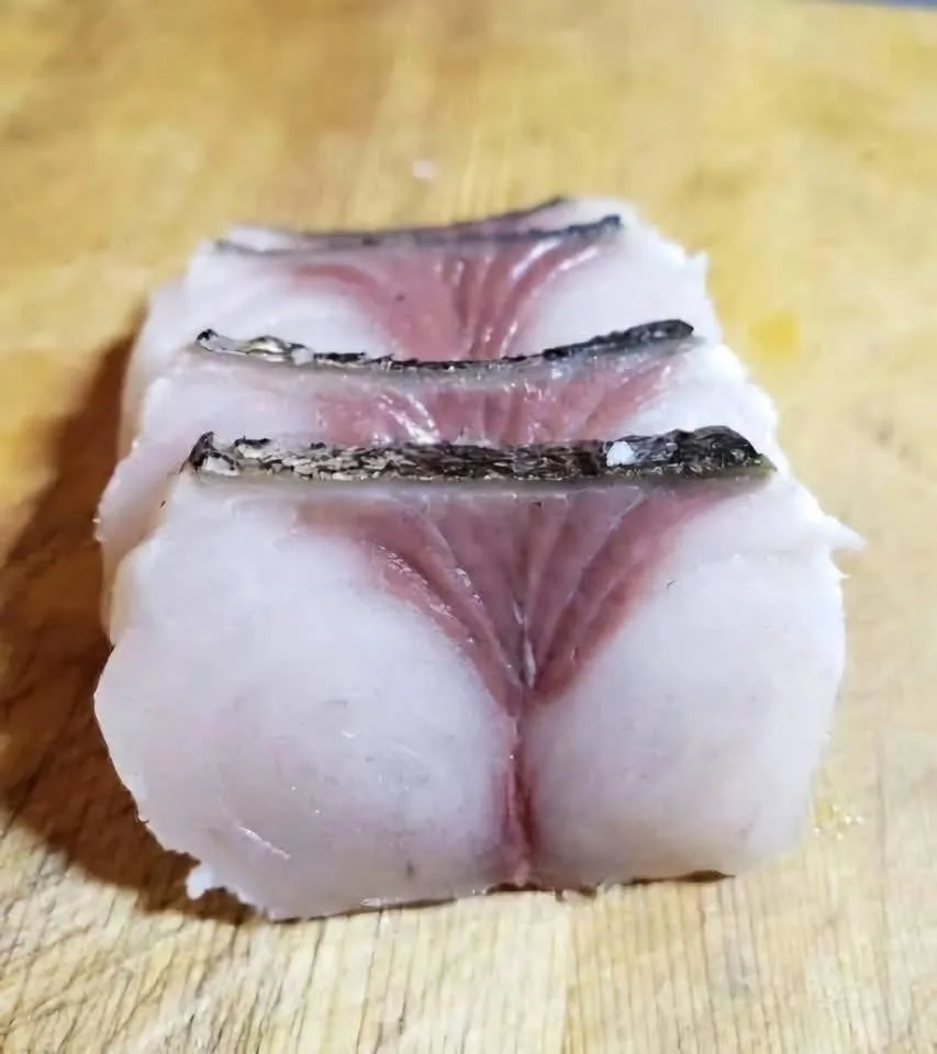 a close-up photo of three stacked slices of raw fish fillet on a wooden cutting board, showing the translucent flesh and…