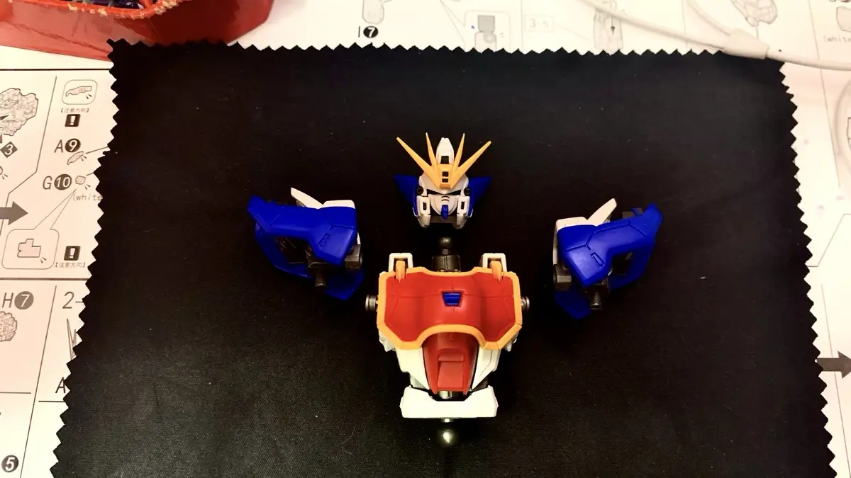 A partially assembled plastic model of a Gundam robot, with its head, torso, and two arms separated and laid out on a black…