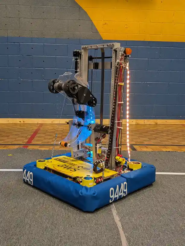 A custom-built robot with a blue padded base and the number 9449 on its sides, featuring a tall metal frame, exposed wiring…