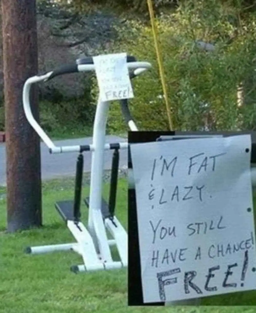 An outdoor exercise machine sits on grass next to a tree, with a handwritten sign attached that reads 'I'M FAT & LAZY. You…