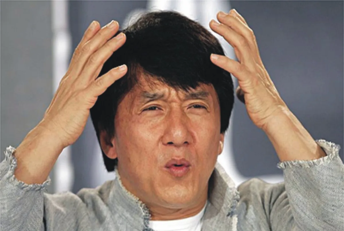 a close-up of actor Jackie Chan with both hands on his head, looking bewildered or exasperated, wearing a grey jacket over a…