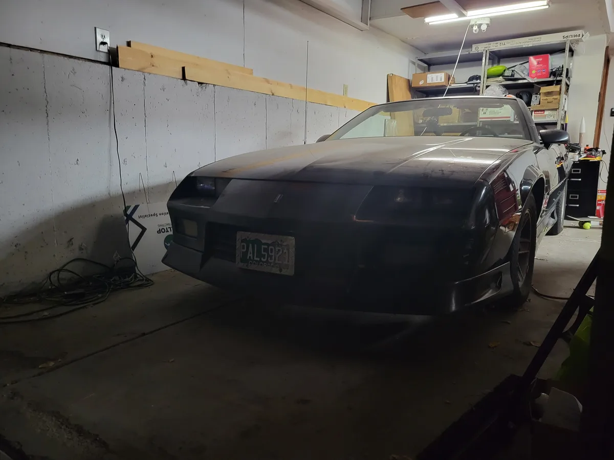 A black Nissan 240SX sports car is parked inside a cluttered garage, its front end slightly dusty and showing signs of…