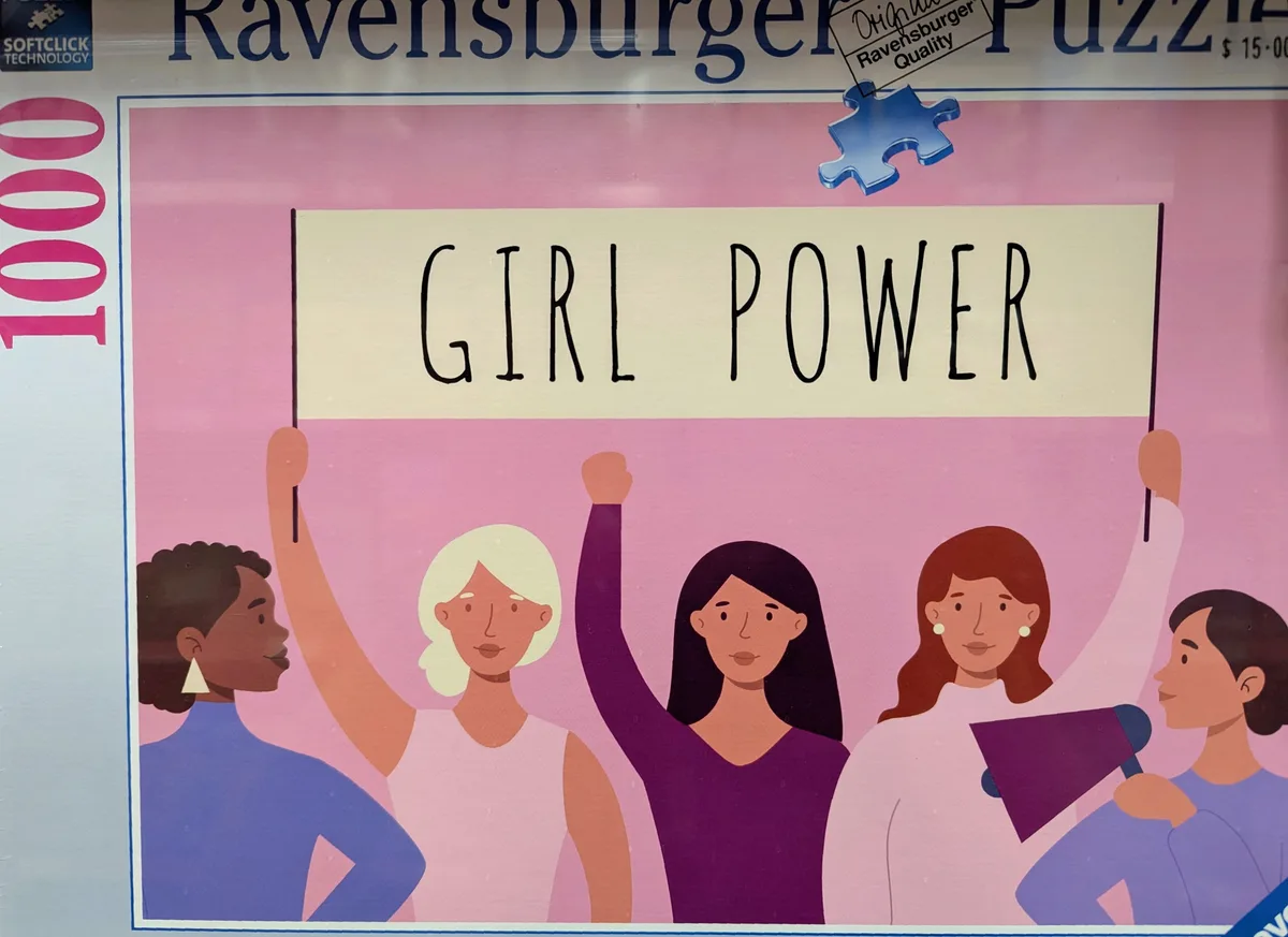 A Ravensburger 1000-piece puzzle box shows a pink-themed illustration of five diverse women holding a sign that reads 'GIRL…