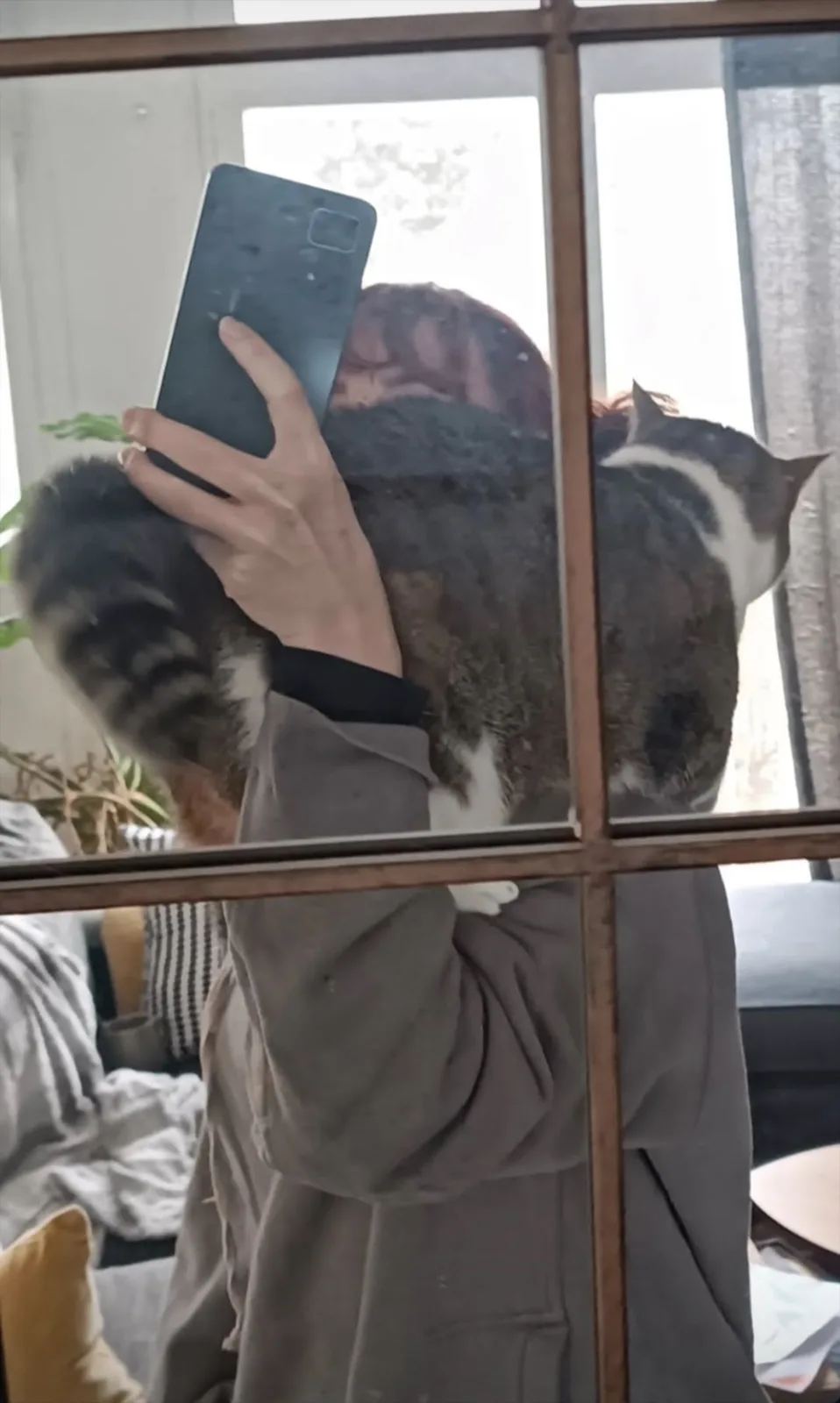 A person in a gray robe is holding a fluffy tabby cat while taking a mirror selfie with a smartphone. The cat is wrapped…