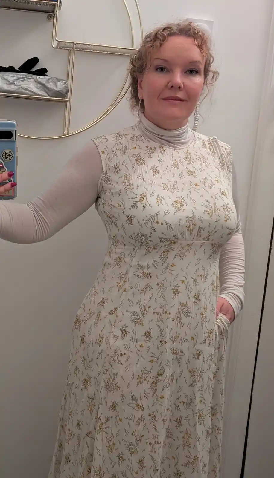 A woman with curly blonde hair takes a mirror selfie wearing a white floral dress over a white turtleneck, holding a phone…