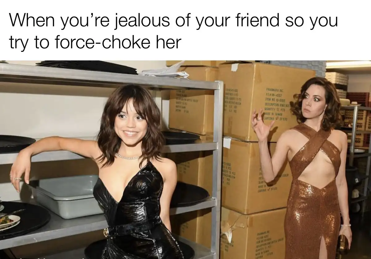 Two women in glamorous dresses stand in a storage room with shelves and cardboard boxes. The woman on the left smiles…