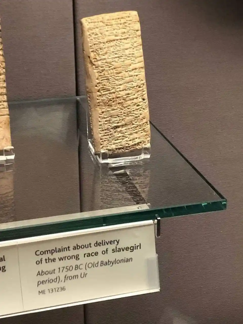 A museum display shows an ancient cuneiform tablet on a glass shelf, with a label reading 'Complaint about delivery of the…