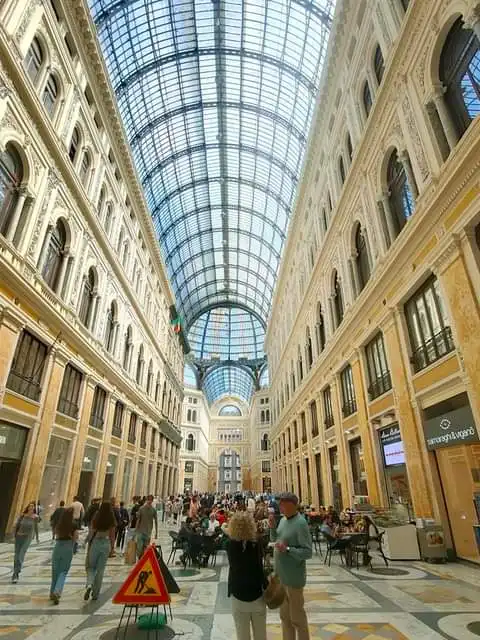 A wide-angle photo of a grand, ornate shopping arcade with a glass and metal vaulted ceiling, bustling with people walking…