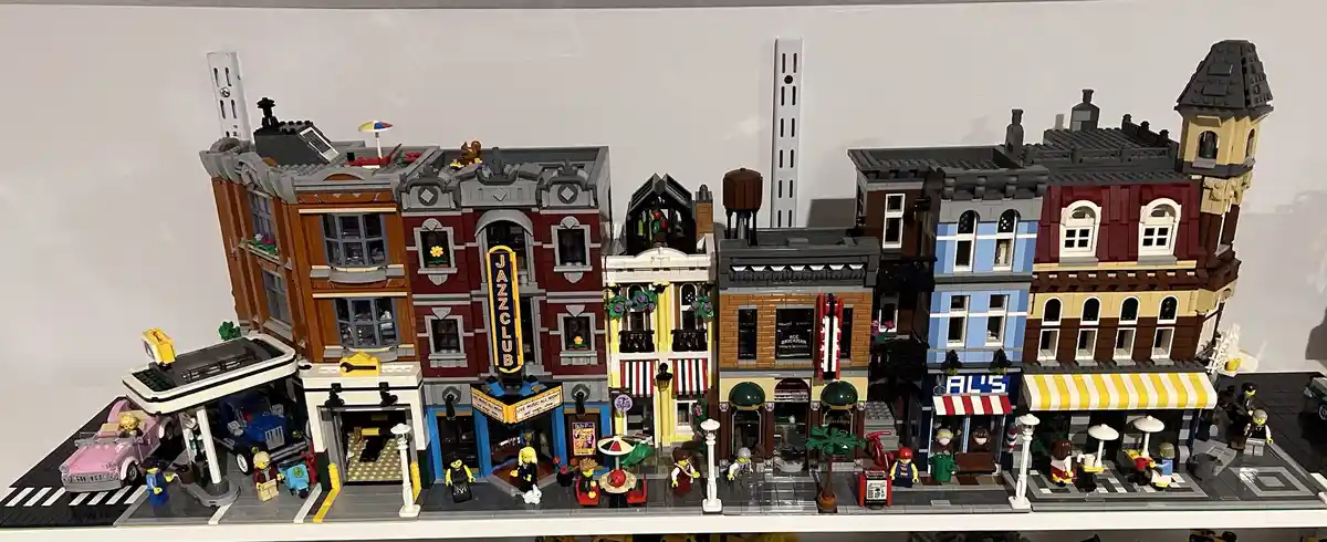 A detailed LEGO city street scene with multiple buildings, including a prominent red brick building with a vertical yellow…