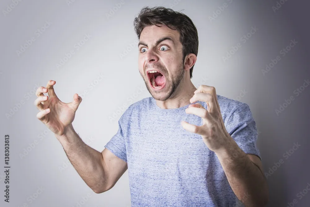a man with dark hair and a beard, wearing a blue heathered t-shirt, is captured mid-expression with wide eyes, an open mouth…