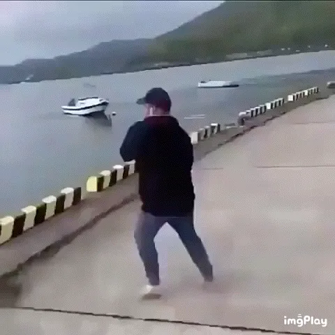 A man on a concrete pier aims a water gun at a boat in the water, then loses his balance and falls dramatically into the…
