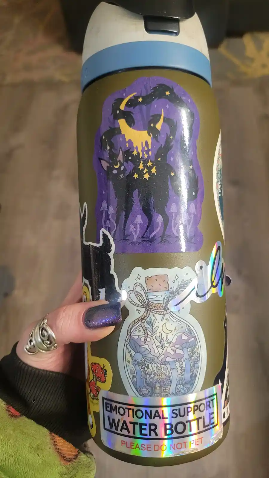 A hand holds a decorated water bottle covered in stickers, including a large purple one with a black cat and crescent moon…