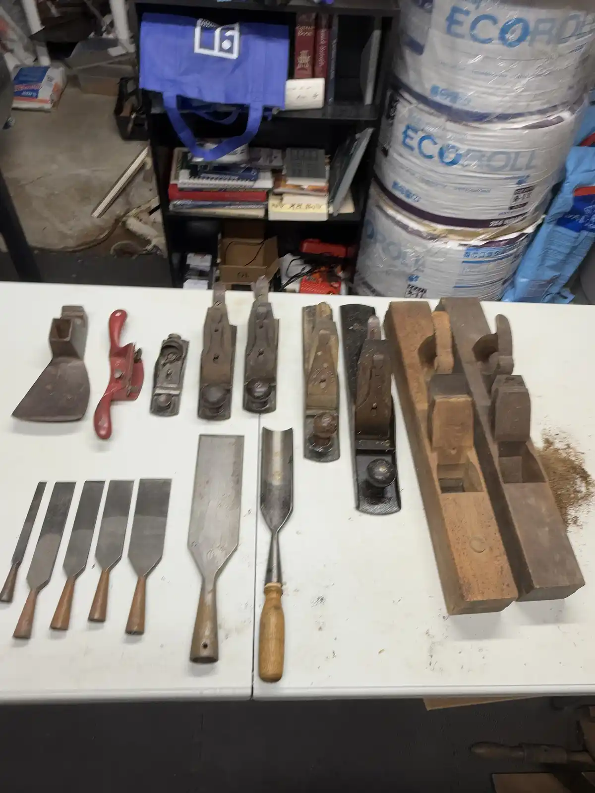 A white table displays a collection of various woodworking tools including chisels, hand planes, and a red hand plane…