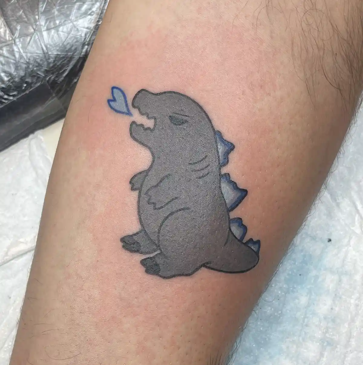 A freshly inked tattoo on a person's leg features a cute, chibi-style Godzilla with a blue heart-shaped breath emanating…