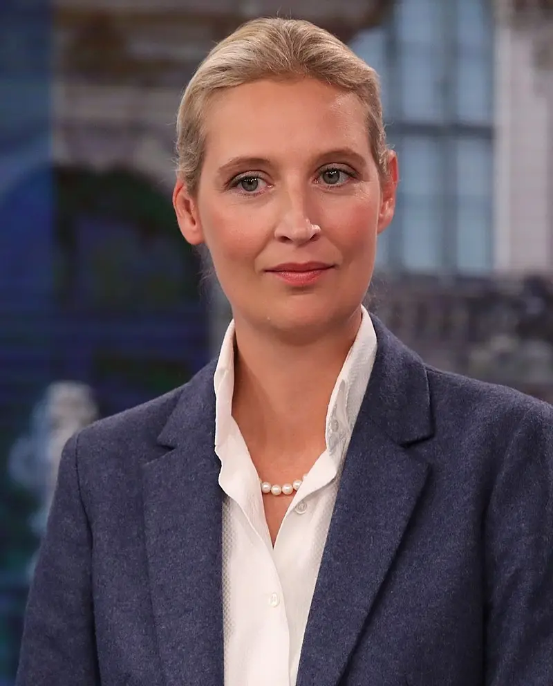 A woman with blonde hair pulled back, wearing a blue blazer over a white collared shirt and a pearl necklace, looks directly…