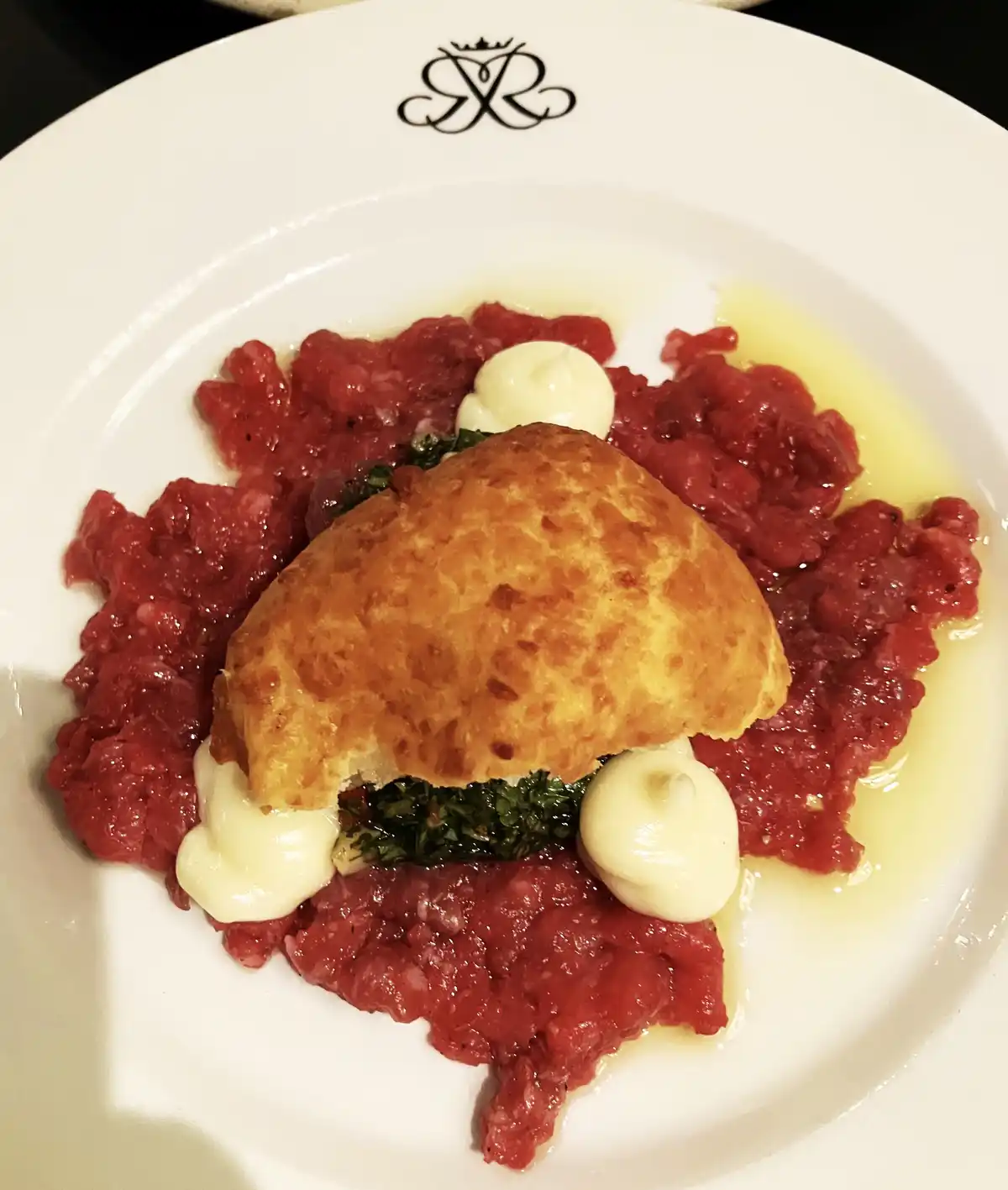 A close-up photo of a gourmet dish on a white plate with a black logo, featuring a golden-brown fried patty-like item…