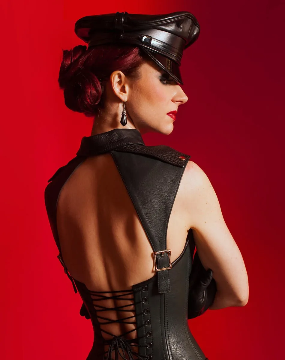 A woman with red hair in a bun, wearing a black leather police-style cap and a revealing black leather corset with lacing at…