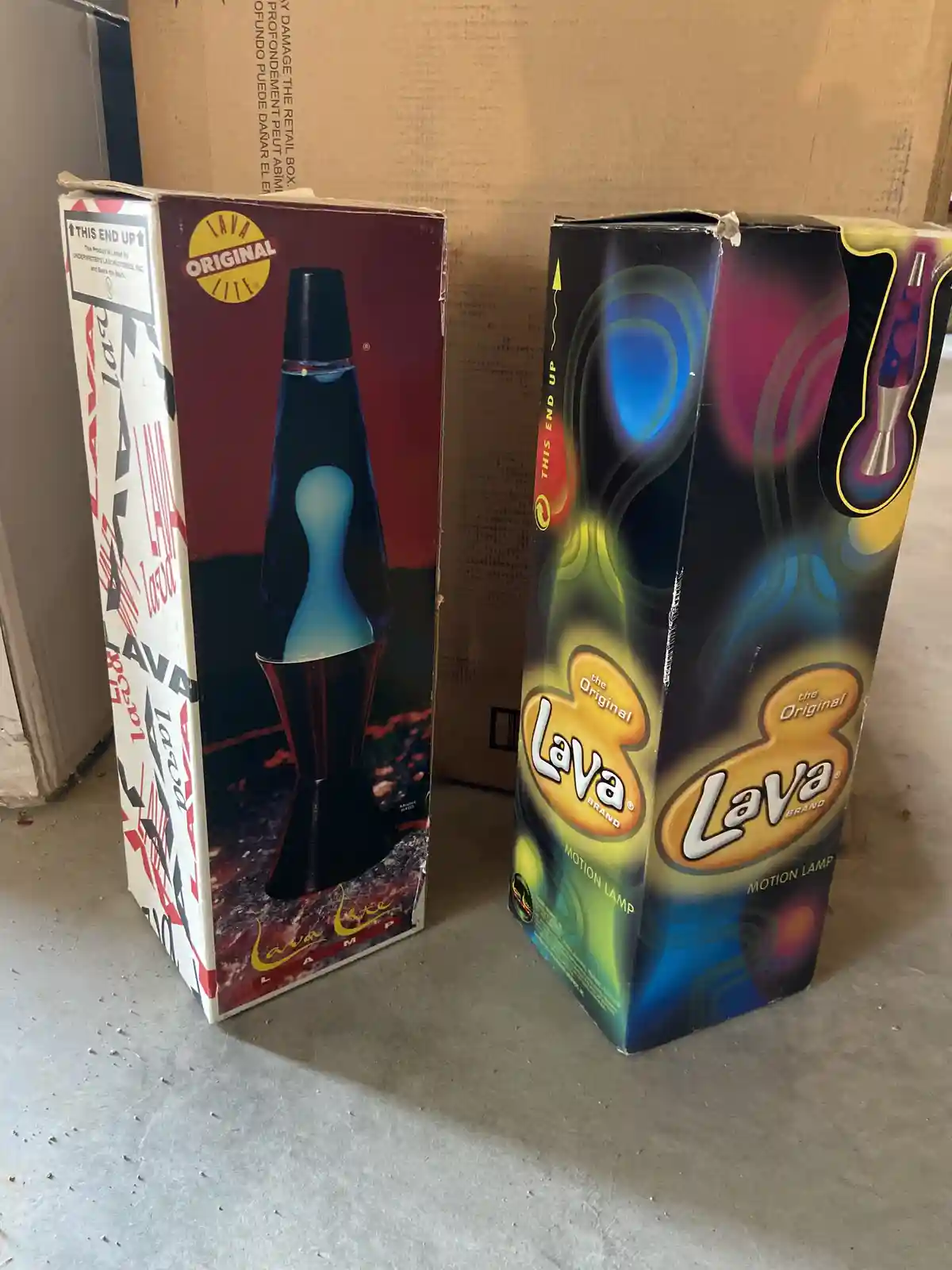 Two vintage Lava Lamp boxes sit side by side on a concrete floor; the left box shows a classic black lava lamp with blue…