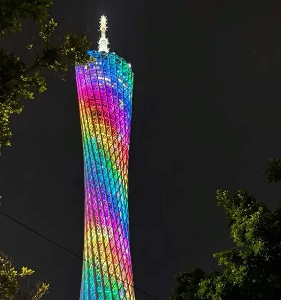 a tall, slender tower with a lattice-like structure is illuminated with a full spectrum of rainbow colors at night, framed…