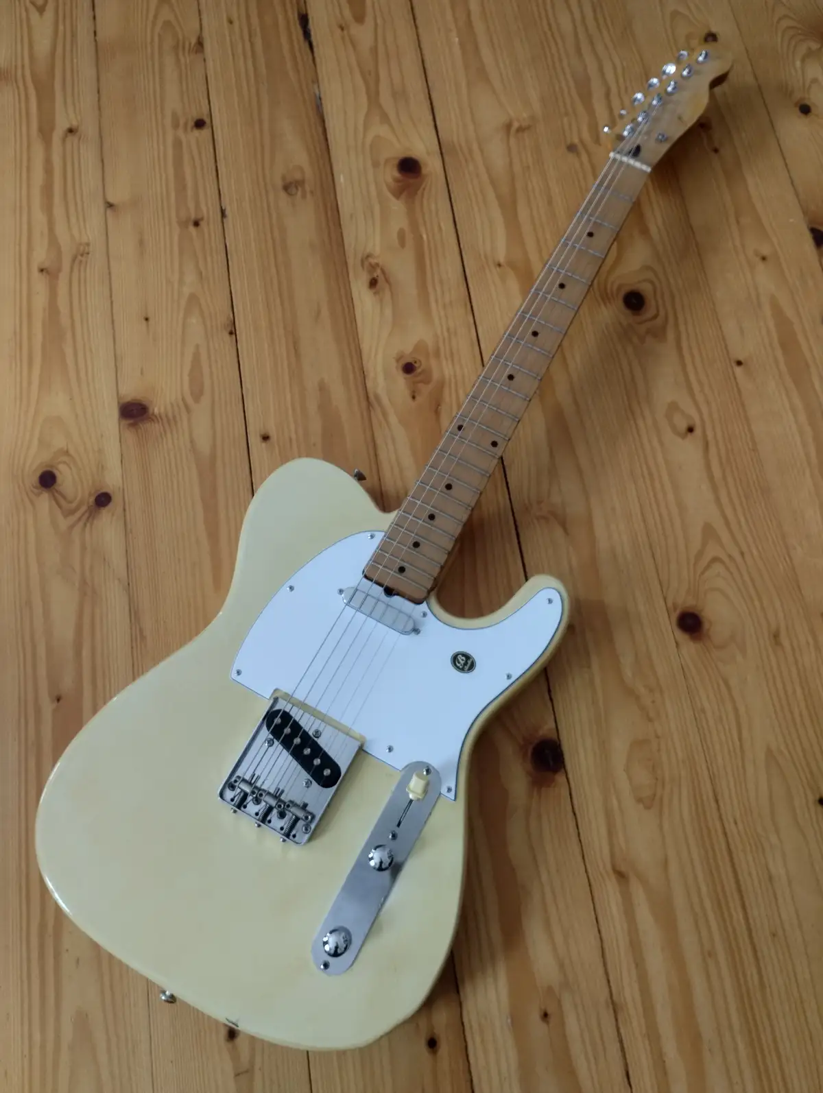 A cream-colored electric guitar with a white pickguard and chrome hardware lies diagonally on a light wood floor. The…