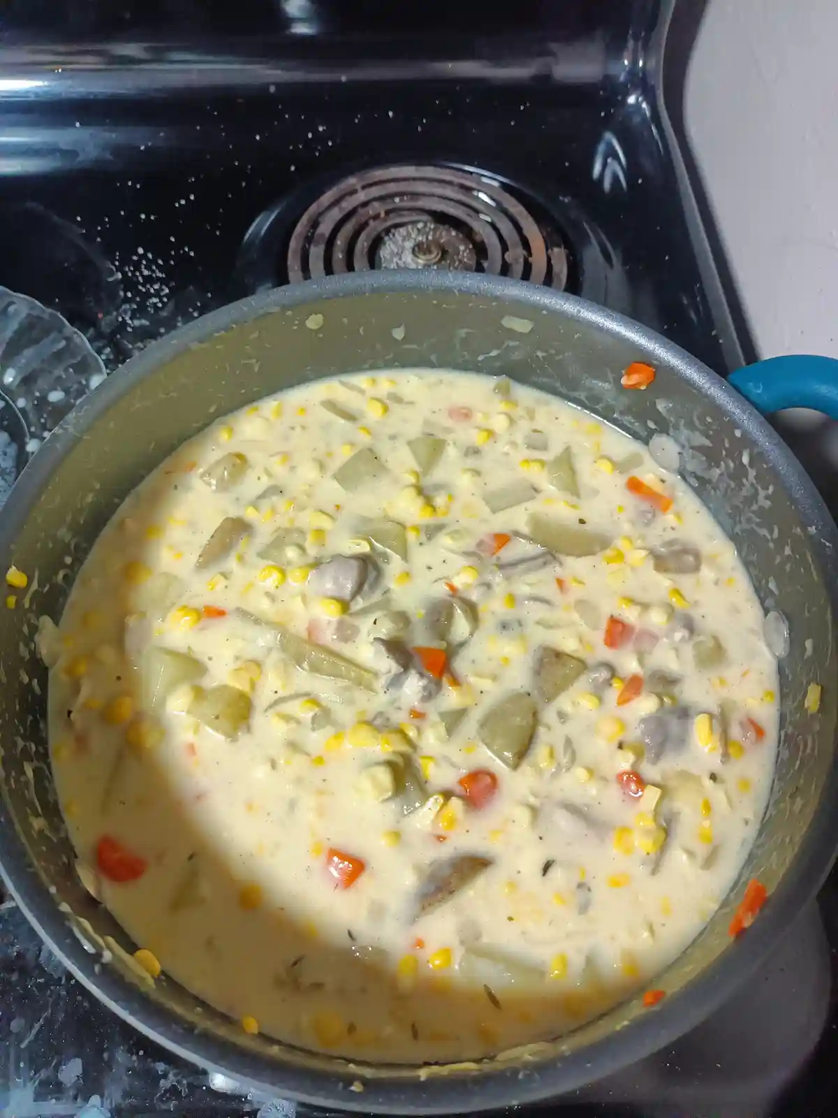 A close-up photo shows a pot of creamy, chunky soup simmering on a black stovetop. The soup contains visible yellow corn…