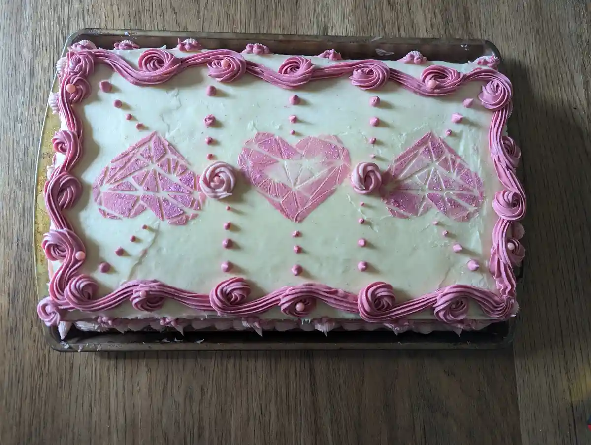 A rectangular cake with white frosting and pink icing decorations sits on a metal baking tray on a wooden surface. The cake…