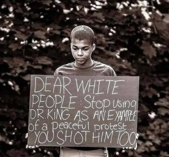 A young Black man stands outdoors holding a handwritten sign that reads 'DEAR WHITE PEOPLE: Stop using DR KING AS AN EXAMPLE…