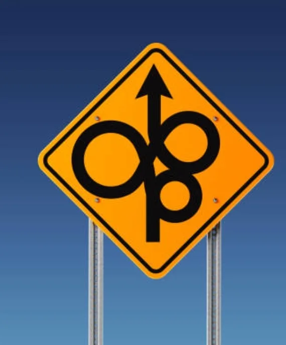 A yellow diamond-shaped road sign with a black arrow pointing up through two interlocking circles, resembling the letter 'O'…