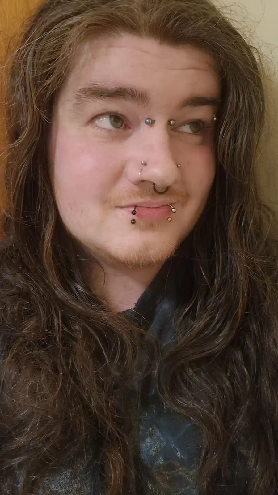 A person with long brown hair and multiple facial piercings, including nose, lip, and eyebrow piercings, looks sideways with…