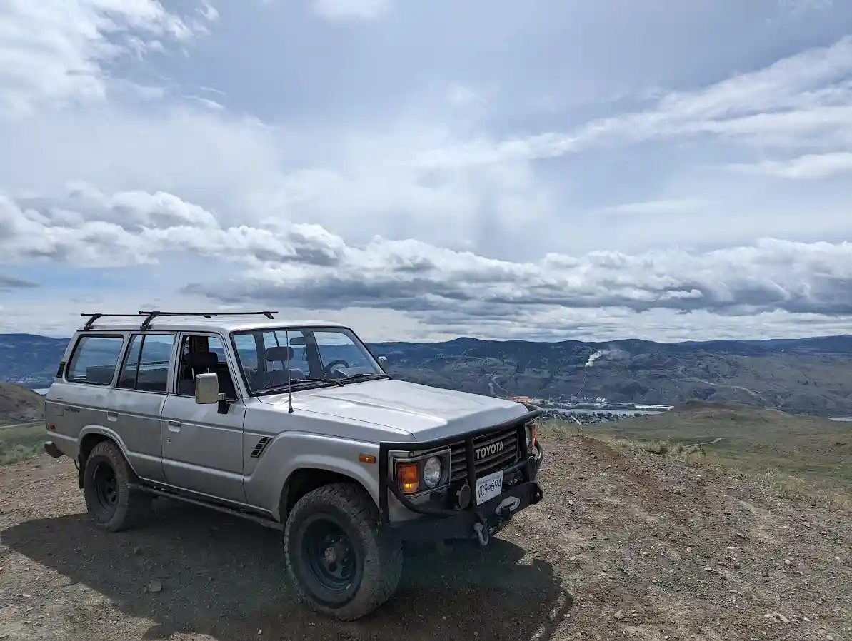 A silver Toyota Land Cruiser SUV is parked on a dirt overlook with a vast mountainous landscape and cloudy sky behind it…