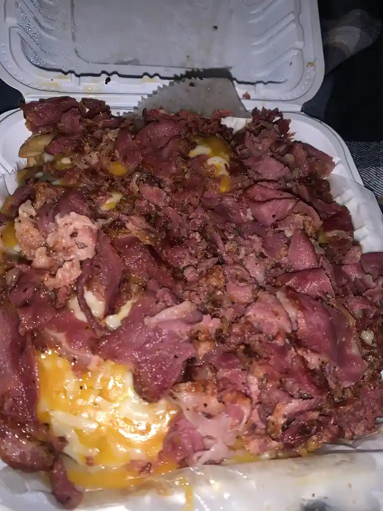 A close-up photo shows a white takeout container filled with a messy, overflowing dish of shredded corned beef, melted…