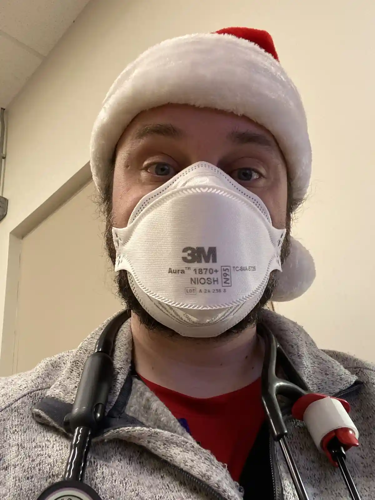 A man wearing a white 3M N95 respirator mask, a red and white Santa hat, and a stethoscope around his neck, looks directly…