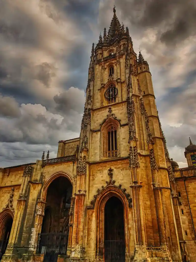 A low-angle photograph of a grand, ornate Gothic cathedral with a tall, intricately carved stone tower under a dramatic…