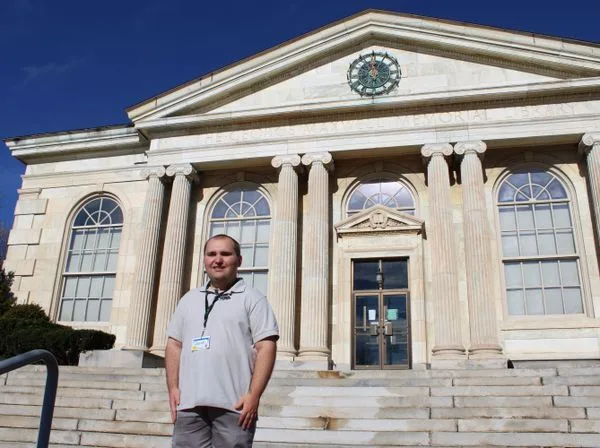 A man in a grey polo shirt and lanyard stands on the steps of a grand neoclassical building with columns and a clock above…