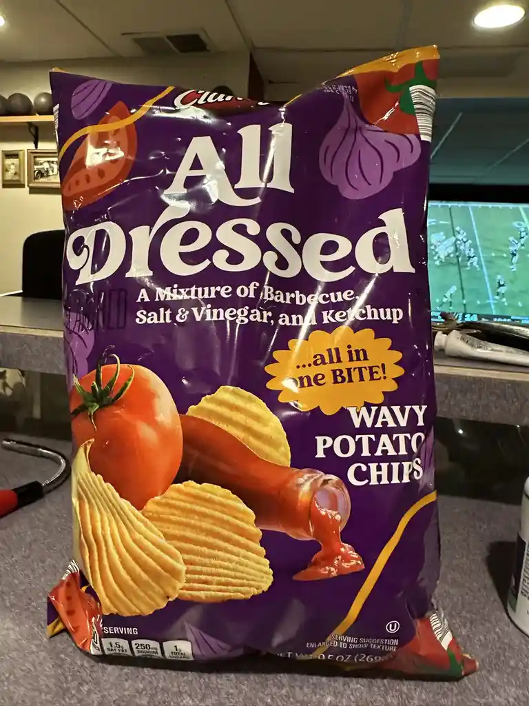 A close-up photo of a purple bag of 'All Dressed' wavy potato chips, which advertises a mixture of barbecue, salt & vinegar…