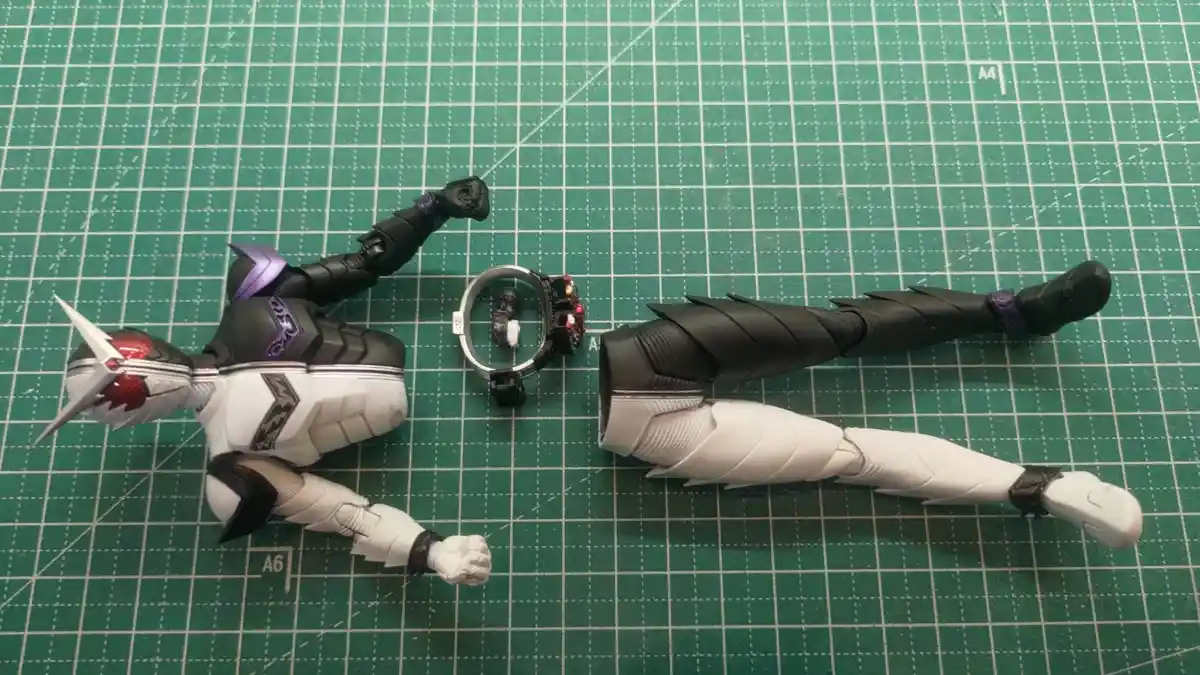 A disassembled Kamen Rider figure lies on a green cutting mat, with its torso, arms, legs, and head separated. The figure's…